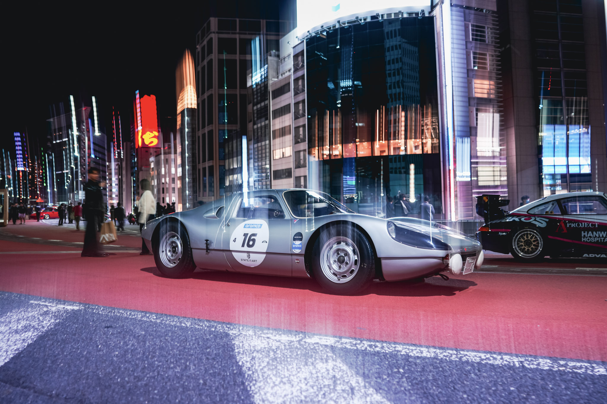 Silver vintage Porsche race car on a neon-lit Tokyo street with light trails and pedestrians